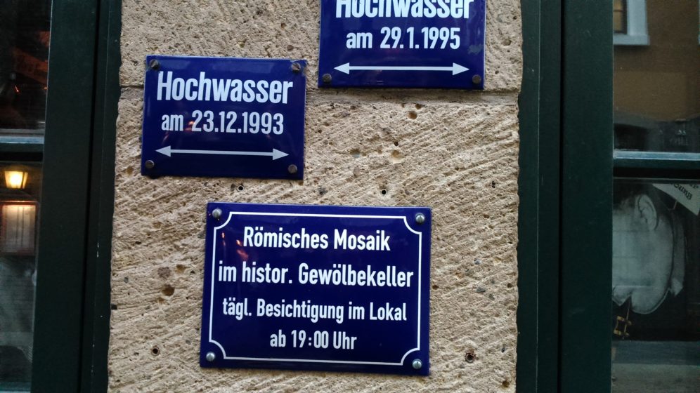 Auf dem Schild steht, dass das römische Mosaik täglich ab 19 Uhr im Lokal besichtigt werden kann. Weitere Schilder weisen auf das Hochwasser in den Jahren 1993 und 1995 hin.