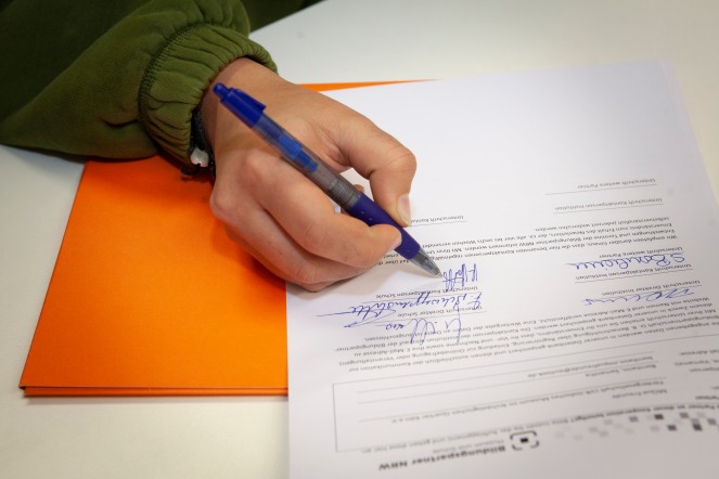 Nahaufnahme von der rechten Hand einer Schülerin, die gerade die Vereinbarung mit einem blauen Kugelschreiber unterzeichnet. Am Bildrand ist noch der Beginn ihres Pullover Ärmels zu sehen.