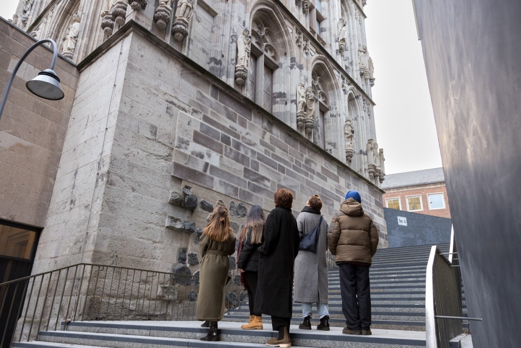 Fünf Frauen in Wintermänteln stehen auf einer Treppe und schauen hoch zu den Steinfiguren auf dem Kölner Rathausturm